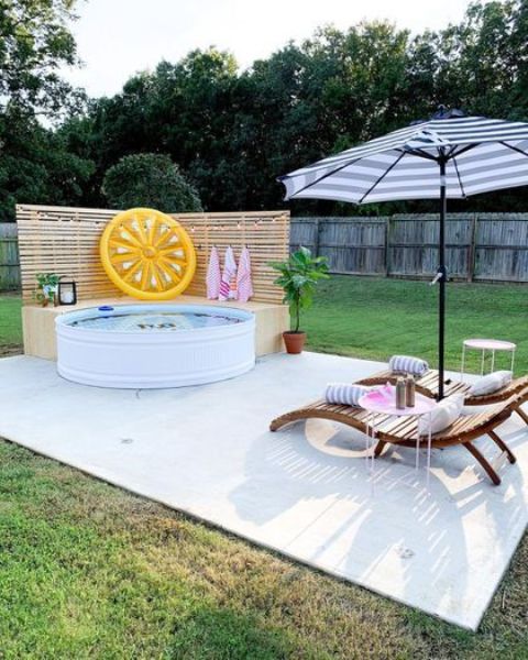 a tiled deck with a stock tank pool, a wooden bench, greenery and a float, loungers and an umbrella