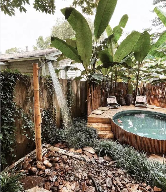 a tropical outdoor space with a lot of greenery and rocks around, a stock tank pool clad with bamboo, with a wooden deck and a bamboo shower