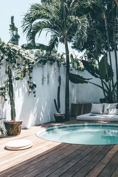a tropical outdoor space with a wooden deck, a daybed with pillows, a stock tank pool and some potted plants and trees