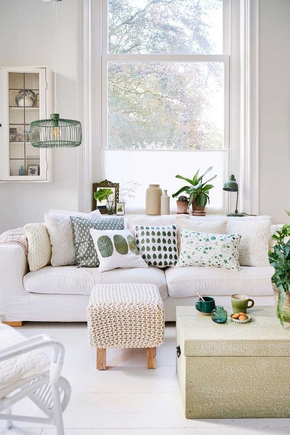 a white living room with a large sofa with printed pillows, a chair, a woven pouf and a coffee table, some cool decor