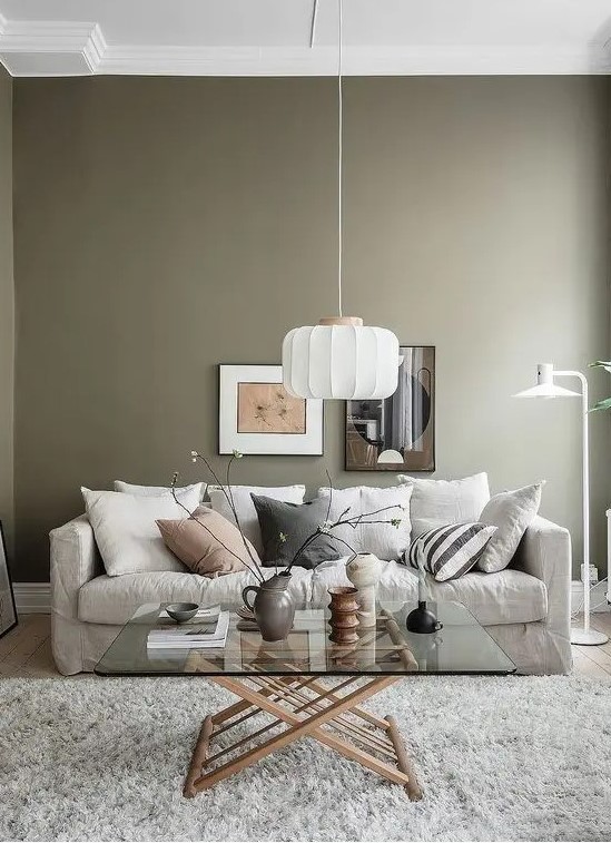 an airy living room with olive green walls, a neutral sofa and pillows, a glass coffee table, a pendant lamp and some art