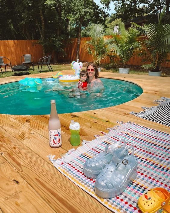 An in ground stock tank pool with a wooden deck, potted plants and trees and some outdoor furniture