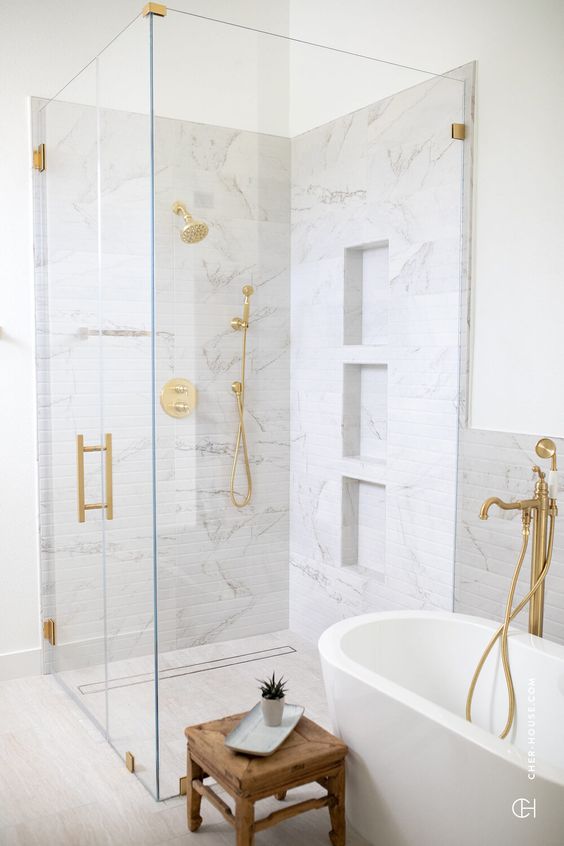 a beautiful neutral bathroom with a shower clad with white marble tiles, an oval tub, gold fixtures, a wooden stool