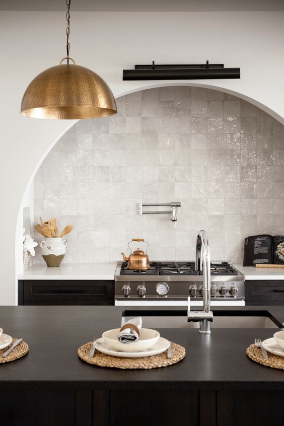 A chic kitchen with a black kitchen island and cabinets, an arched niche over the cooker with a built in hood and white Zellige tiles