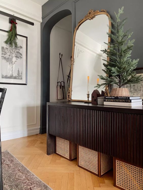 a beautiful dark-stained reeded console table with cane boxes under it, some coffee table books, an arched mirror and a Christmas tree