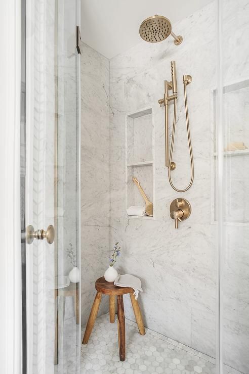 a chic shower space done with marble tiles, niches used for storage, brass fixtures and a wooden stool