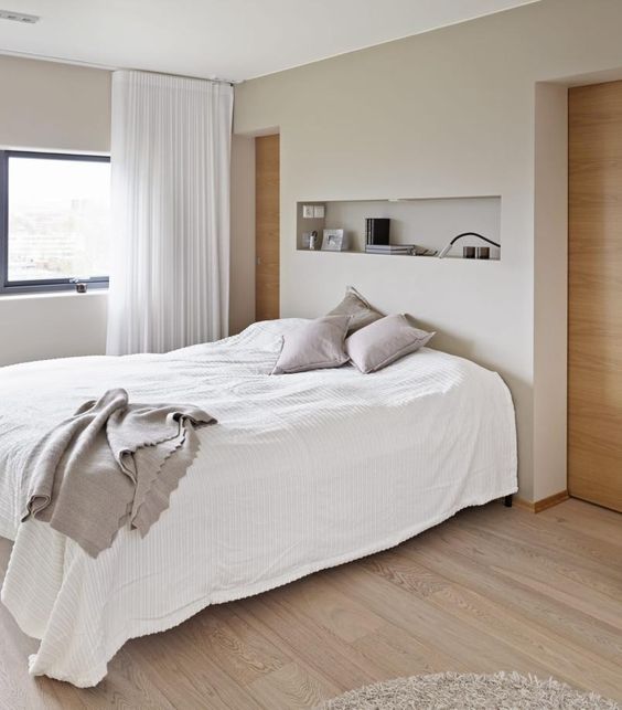 a neutral bedroom with a bed and neutral bedding, a niche used for storage and siplaying, a small window and neutral curtains