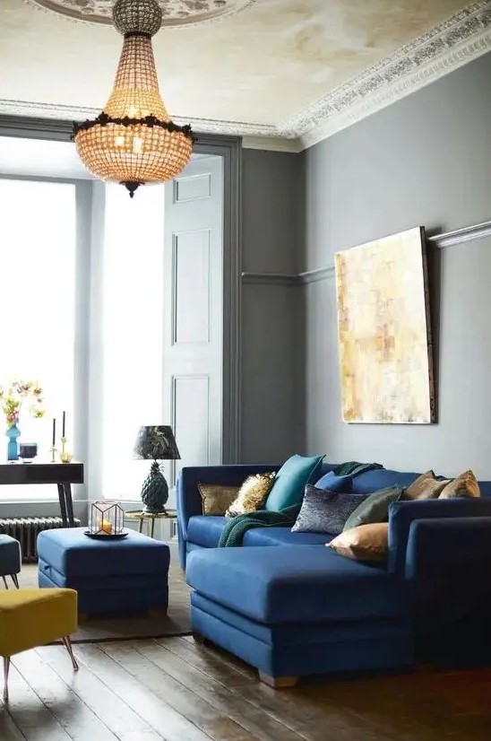 A beautiful and refined living room with grey walls, a navy mid century modern sectional, a navy pouf and yellow and blue stools, a crystal chandelier and a statement artwork