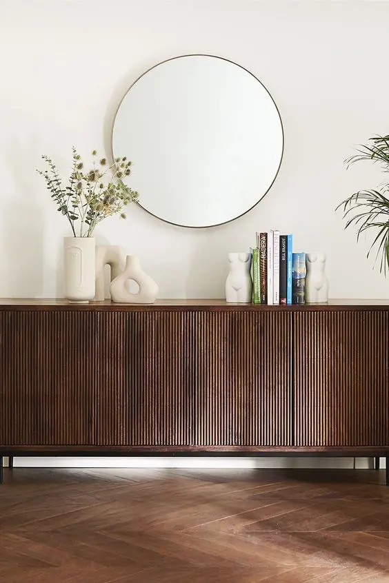 A dark stained sideboard with beautiful decor, vases, books and a round mirror over it is a stylish and catchy idea