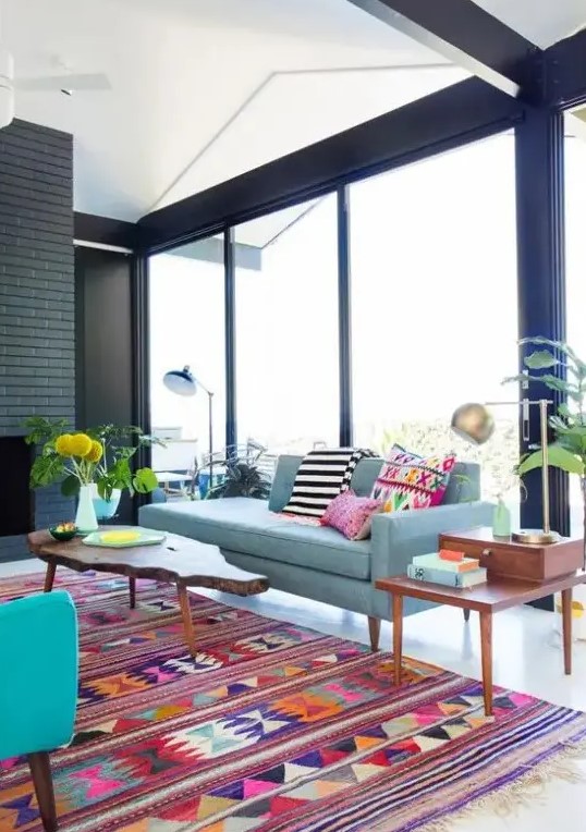 a mid-century modern to boho living room with a black brick fireplace, a blue sofa, a turquoise chair, bright pillows and rugs, some potted plants