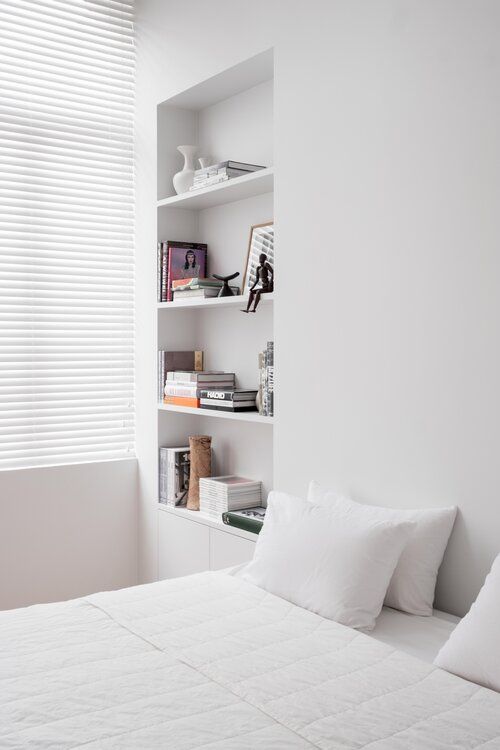 A minimalist white bedroom with a bed and white bedding, a large niche with shelves that are used for decorating and a built in cabinet