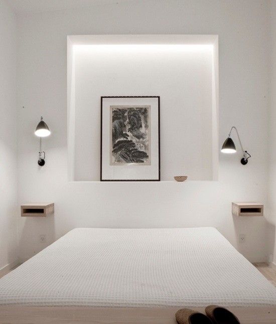 a minimalist white bedroom with a low bed, a niche used for decor, floating mini nightstands and black sconces