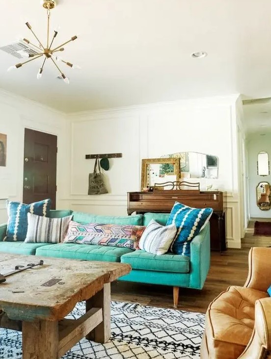 An eclectic living room with a mid century modern turquoise sofa, a rough wooden table, a chair, a wooden dresser and some mirrors on it