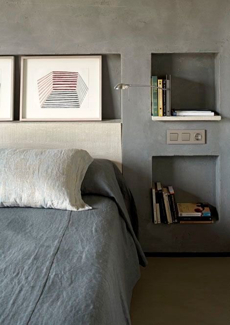 a modern bedroom with concrete walls and niches as nightstands, a sconce and some artwork is a loacnic and edgy space