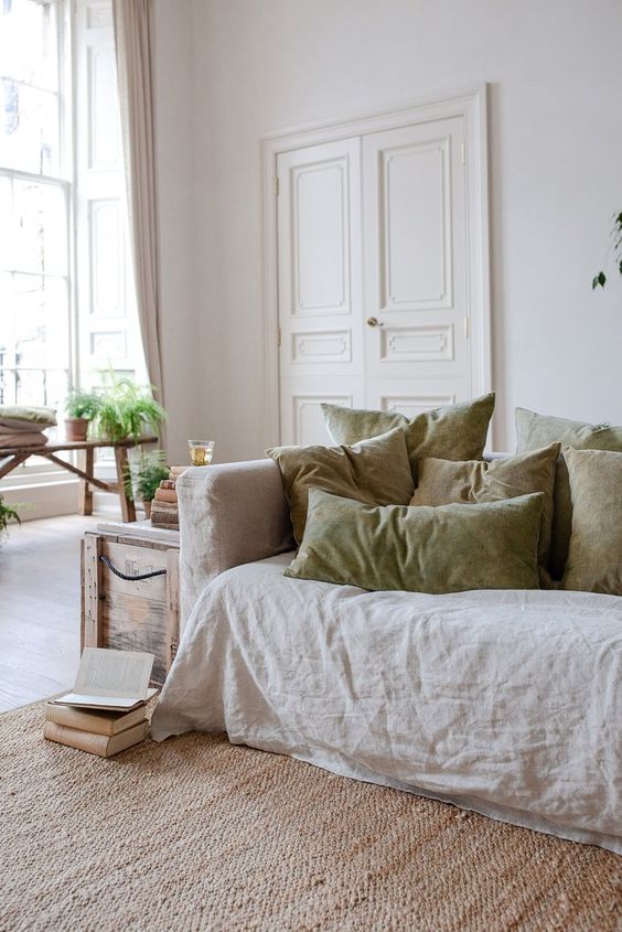 a neutral living room with a linen sofa and matching green linen pillows, a chest, a bench with greenery and a jute rug