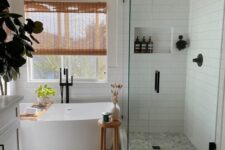 23 a modern farmhouse bathroom clad with grey square and white skinny tiles, a shower space, an oval tub, a white vanity, wooden rugs and shades, a niche in the shower