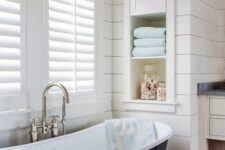 25 a modern farmhouse bathroom done with shiplap, with an oval tub, a window with shutters, a niche with shelves used for decor and towels