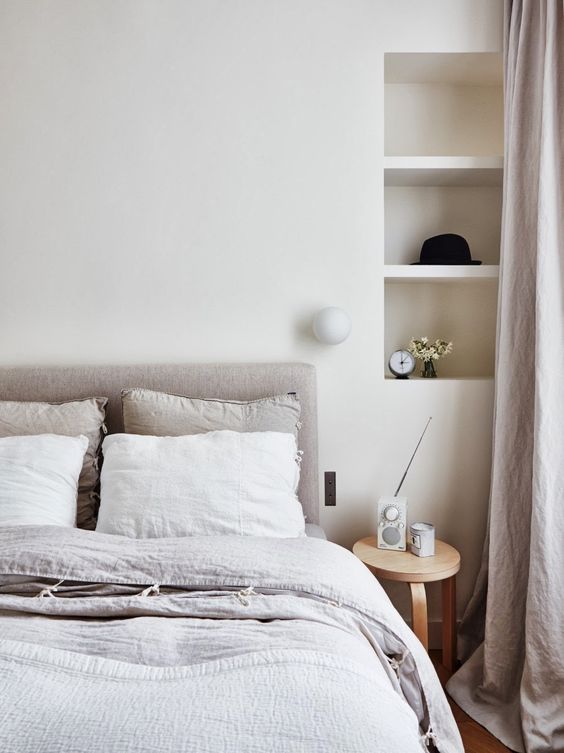 a neutral bedroom with niche shelves, a grey upholstered bed with neutral bedding, a stool as a nightstand feels very soothing