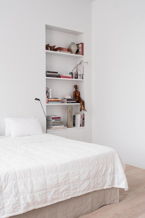 a Scandinavian bedroom with a bed and neutral bedding, a niche with shelves used for storing books and various decor is a lovely space