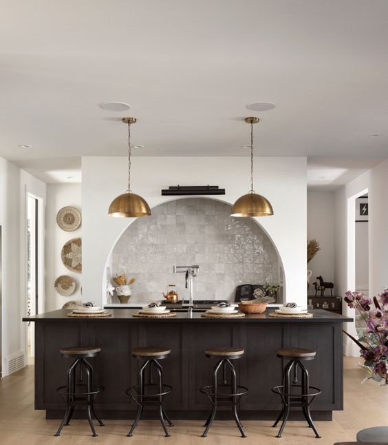 A modern kitchen with an arched niche clad with Zellige tiles and built in cabinets, a black kitchen island, brass pendant lamps and industrial stools