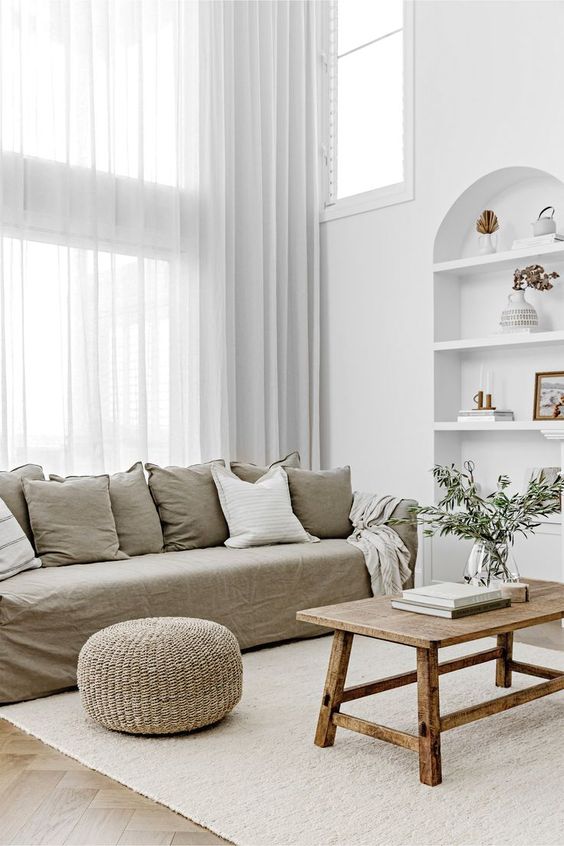 a neutral living room with a grey linen sofa and pillows, a stained table, a niche with shelves, a jute pouf and greenery
