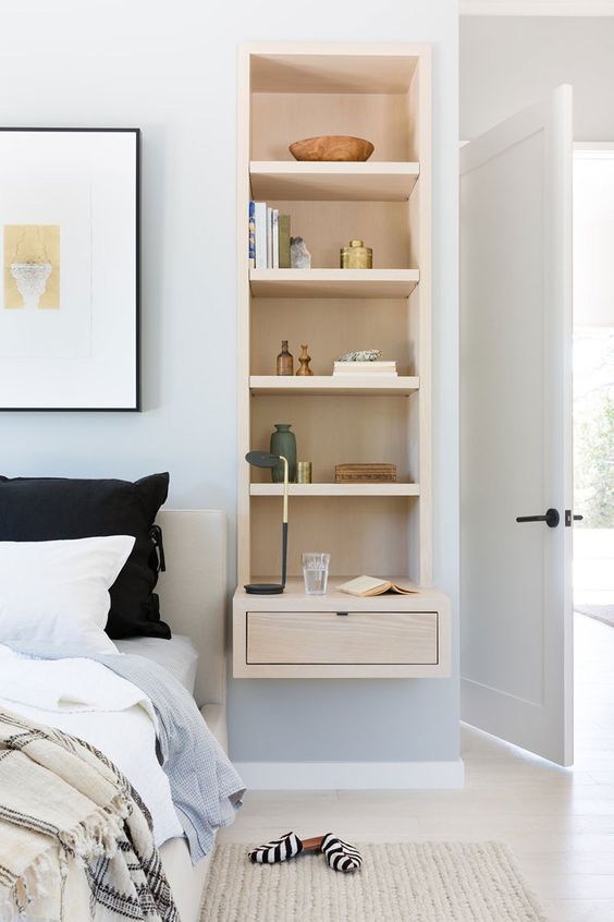 A serene neutral bedroom with pale blue walls, a bed and a niche with a built in storage piece and a drawer, some decor and necessary stuff