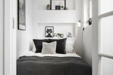 30 a tiny bedroom in white, with only a bed squuzed in, black and white bedding, niche shelves with decor, an artwork and a sconce