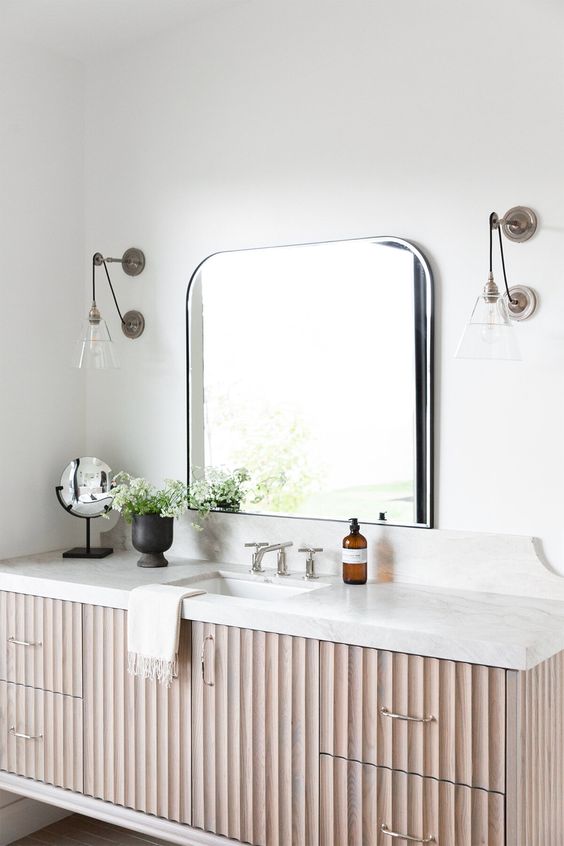 a fluted vanity with stainless steel handles, a white stone countertop, an arched mirror and wall sconces
