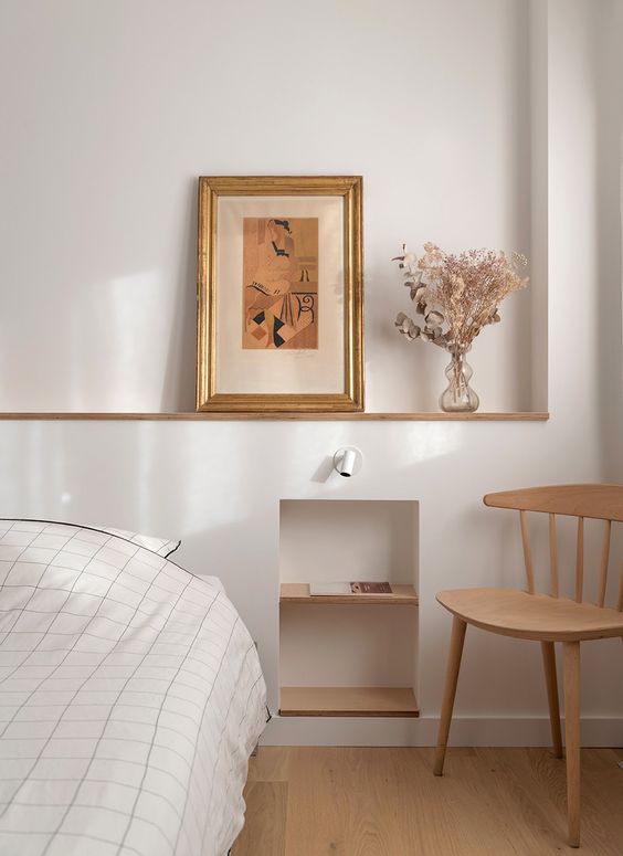 a welcoming neutral bedroom with a bed and neutral bedding, a niche with shelves, some art and a stained chair