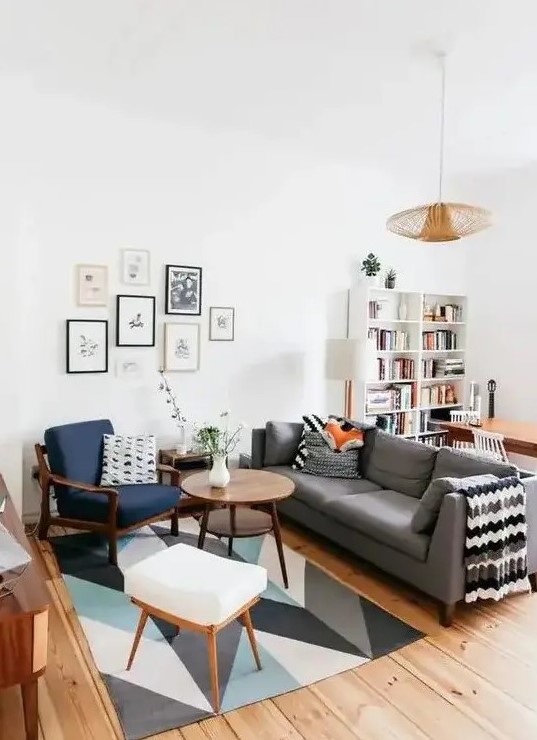 A mid century modern living room with a bold printed rug, a grey sofa, a navy chair and a white stool, a cool gallery wall