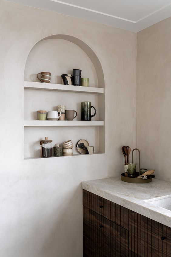 A contemporary kitchen with plaster walls, dark stained cabines and neutral stone countertops, an arched niche for displaying mugs
