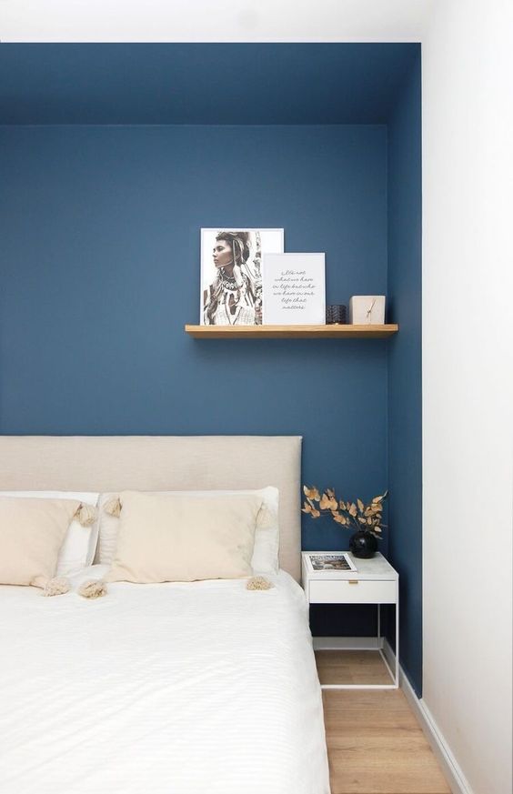 a modern bedroom with a large niche painted navy, a neutral bed with bedding in it, a shelf and a white nightstand