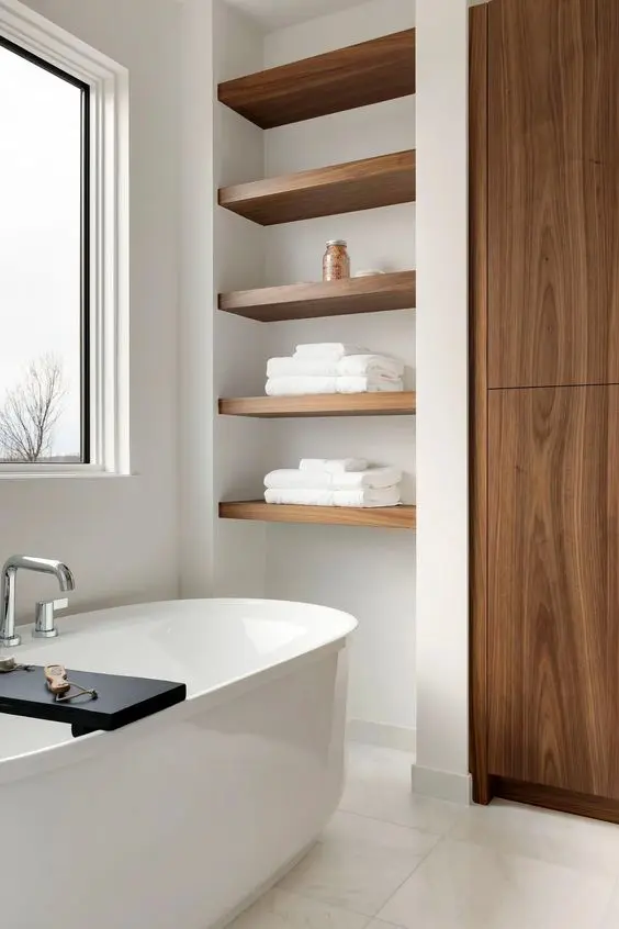A refined contemporary bathroom clad with neutral tiles, a wardrobe, built in shelves in a niche, an oval tub and a window