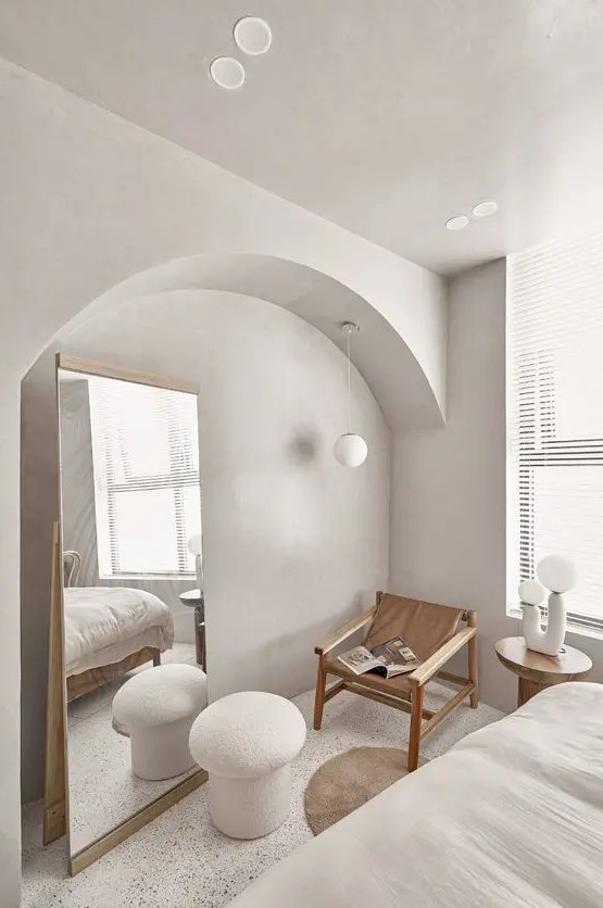 a contemporary bedroom in cold neutral tones, with an arched niche with a floor mirror, a wood and leather chair and a lamp