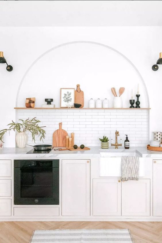 a modern kitchen with neutral lower cabinets, white marble countertops, an arched niche with a tile backsplash and an open shelf used for decor