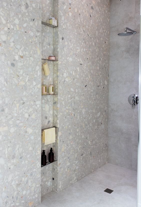a shower space done with concrete tiles and terrazzo, with a tall and narrow niche with shelves used for storing stuff