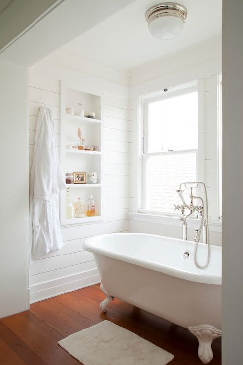 a white farmhouse bathroom clad with shiplap, with a niche with shelves and some lovely decor, a clawfoot tub and windows