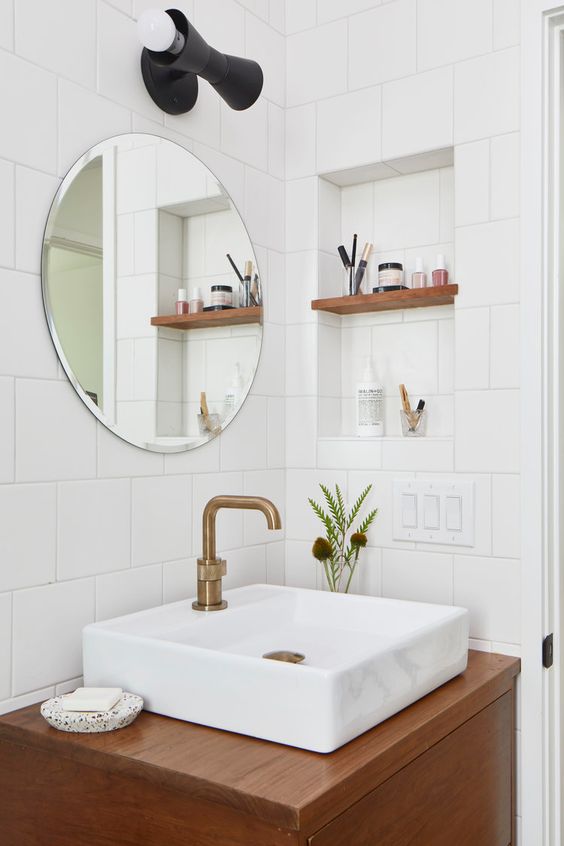 a chic modern powder room with white square tiles, a niche with shelves for storing small things. a stained vanity with a sink and a round mirror