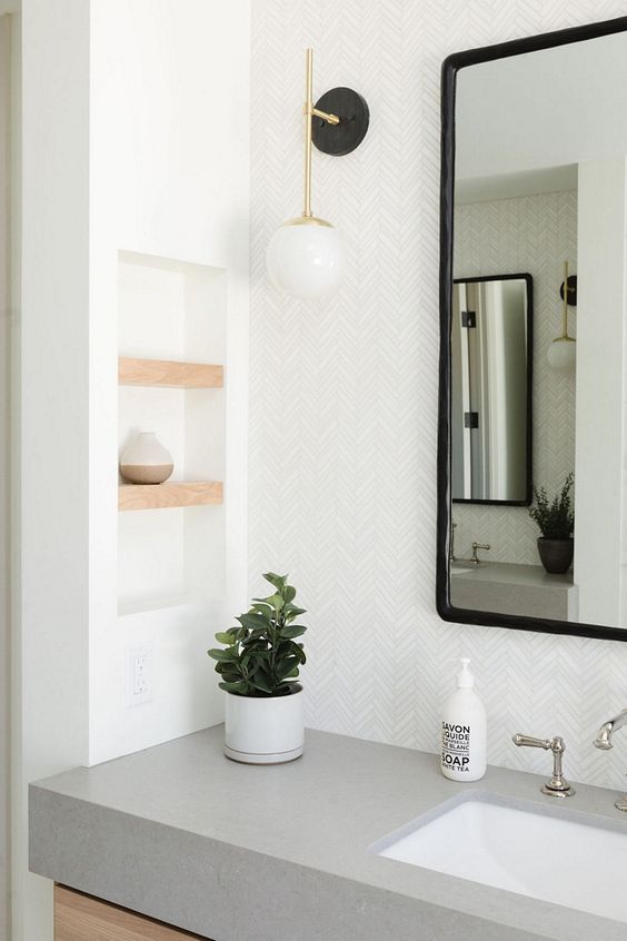 a modern bathroom with herringbone tiles, a cocnrete vanity with a sink, a mirror, a niche with little shelves for decor
