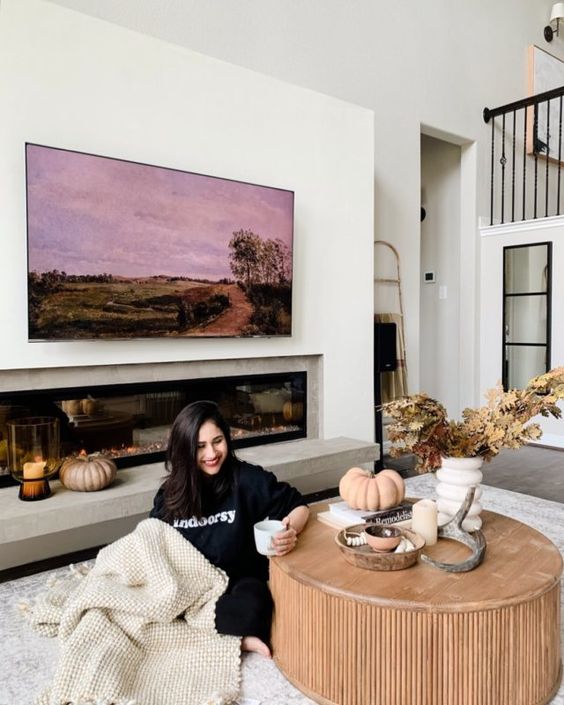 A welcoming living room with a built in fireplace, a large artwork, a round fluted coffee table, a neutral rug and some leaves
