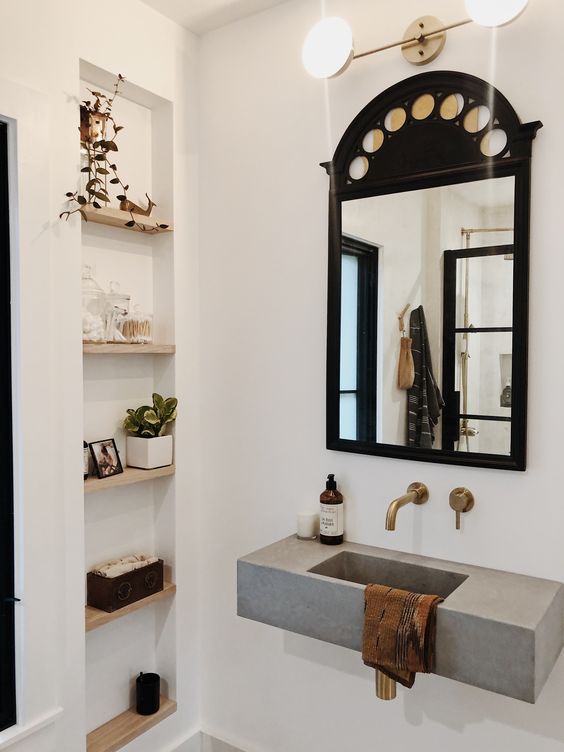 A modern neutral bathroom with a niche with shelves used for decor, a concrete wall mounted sink, a black framed mirror