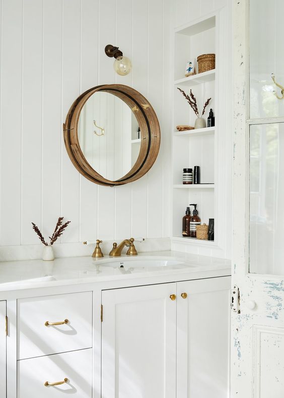 a white farmhouse bathroom clad with skinny tiles, with a large vanity, a niche with shelves to store mostly decor, a round mirror