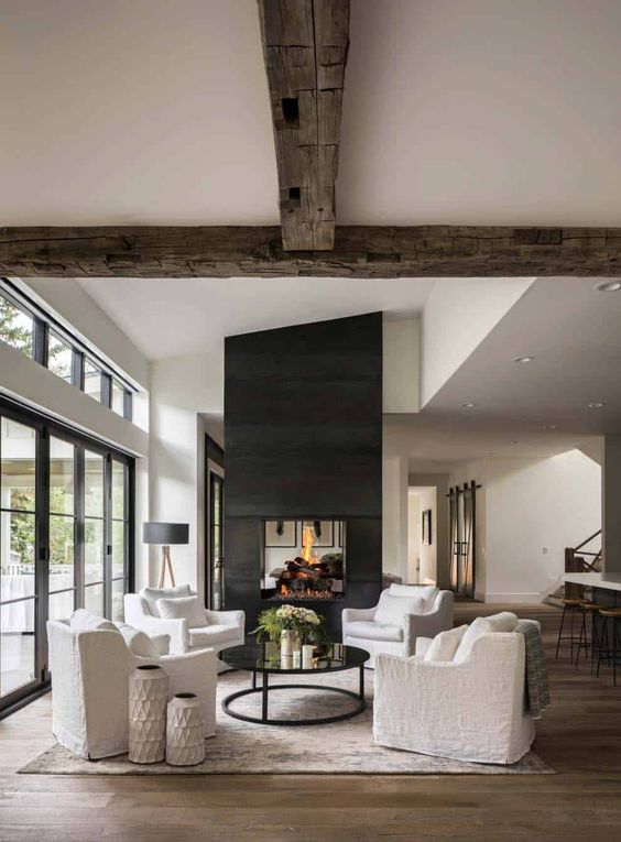 A beautiful modern farmhouse living room with reclaimed wooden beams, a large double sided fireplace, white seating furniture and a coffee table