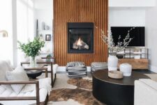 a beautiful modern living room with a fireplace and a reeded surround, white chairs, a black coffee table, printed poufs and a cowhide rug