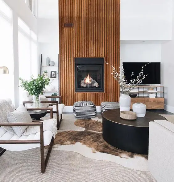 a beautiful modern living room with a fireplace and a reeded surround, white chairs, a black coffee table, printed poufs and a cowhide rug