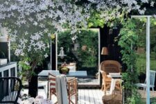a blooming modern farmhouse terrace with a farmhouse feel, rattan and wooden furniture, some pendant lamps and candle lanterns and lots of blooms and greenery