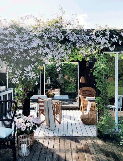 a blooming modern farmhouse terrace with a farmhouse feel, rattan and wooden furniture, some pendant lamps and candle lanterns and lots of blooms and greenery