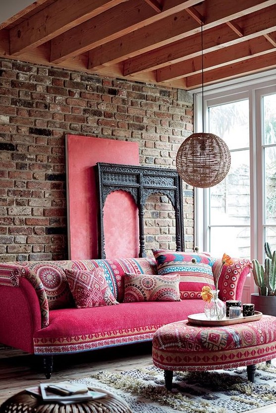 a bright boho living room with a brick wall, a hot pink sofa and ottoman, a wicker lamp and a glazed wall