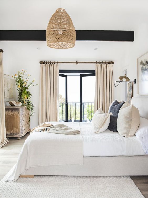 a chic modern farmhouse bedroom with dark-stained wooden beams, an upholstered bed with neutral bedding, a woven pendant lamp and a shabby chic dresser