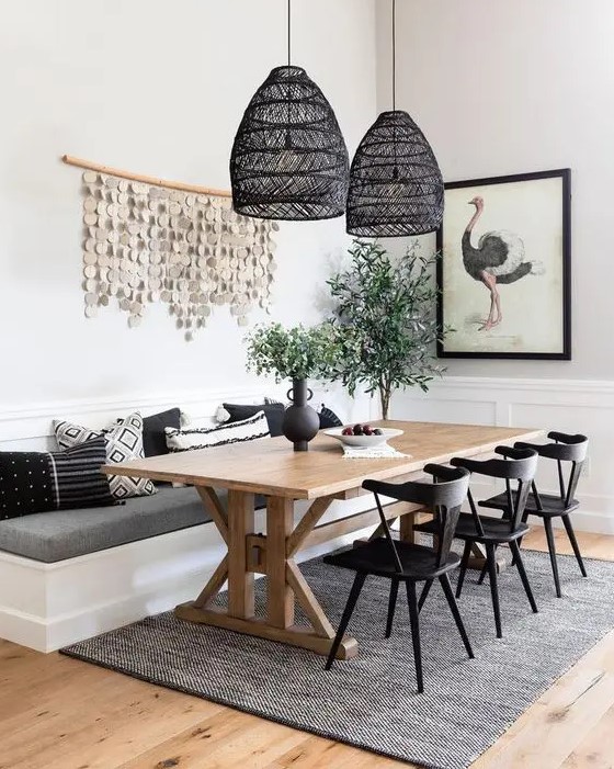 A chic modern farmhouse dining room with white paneled walls, a built in banquette seating, a trestle dining table, black chairs and black woven pendant lamps